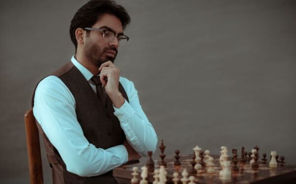 Concentrated bearded male in formal clothes touching chin while sitting on chair and playing chess against gray wall Concentrated bearded male in formal clothes touching chin while sitting on chair and playing chess against gray wall