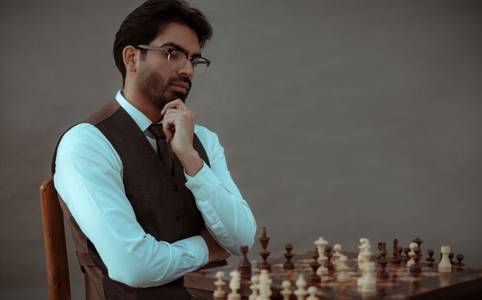 Concentrated bearded male in formal clothes touching chin while sitting on chair and playing chess against gray wall