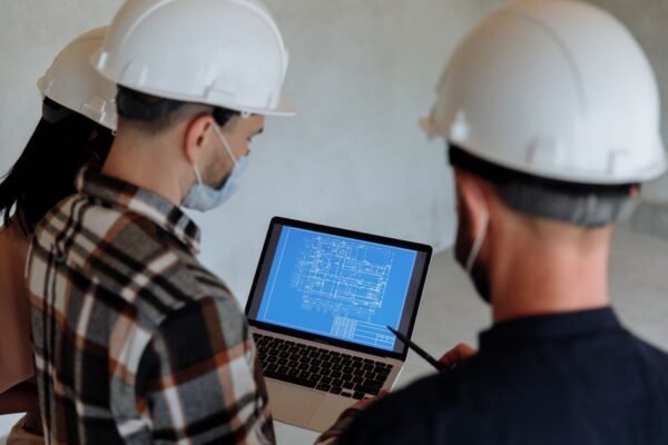 Engineers wearing hard hats and masks review a construction blueprint on a laptop indoors. Engineers wearing hard hats and masks review a construction blueprint on a laptop indoors.